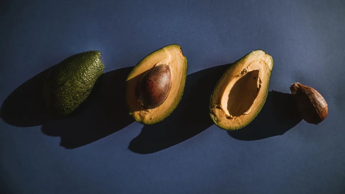 Overhead view of fresh avocados — halved and whole — on a dark blue background. Decorative stock image.