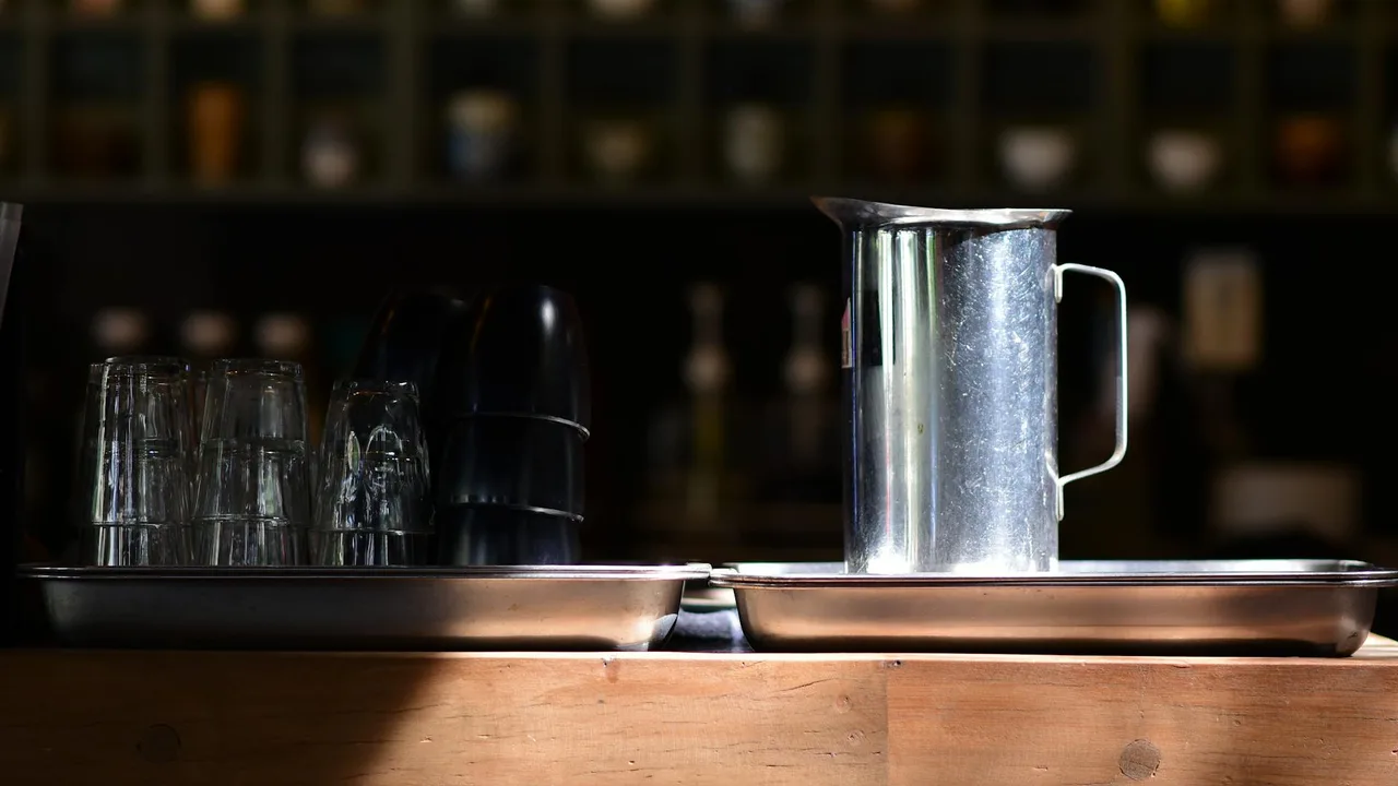 A stainless steel pitcher with glassware on a cafe counter, soft light ambient.