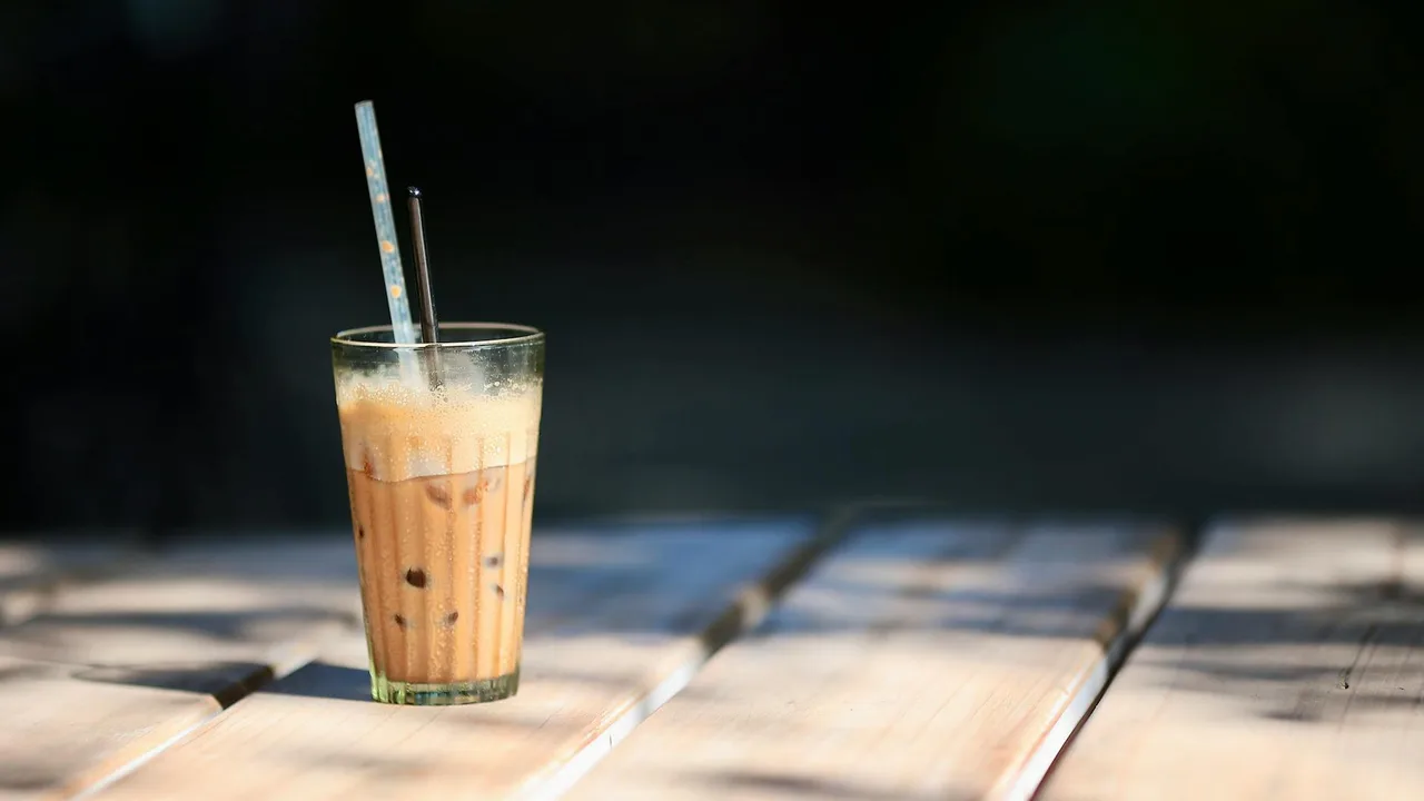 A glass of iced coffee with two straws sits on a wooden table in the sun.