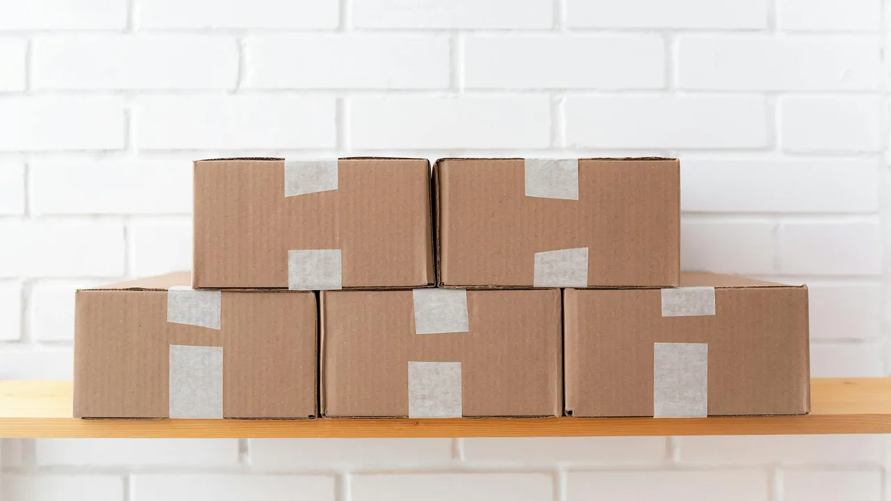 A neat stack of cardboard boxes taped and placed on a wooden shelf against a white brick wall.