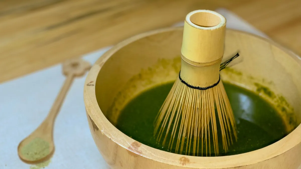 A bamboo whisk in a wooden bowl with matcha tea on a wooden surface.
